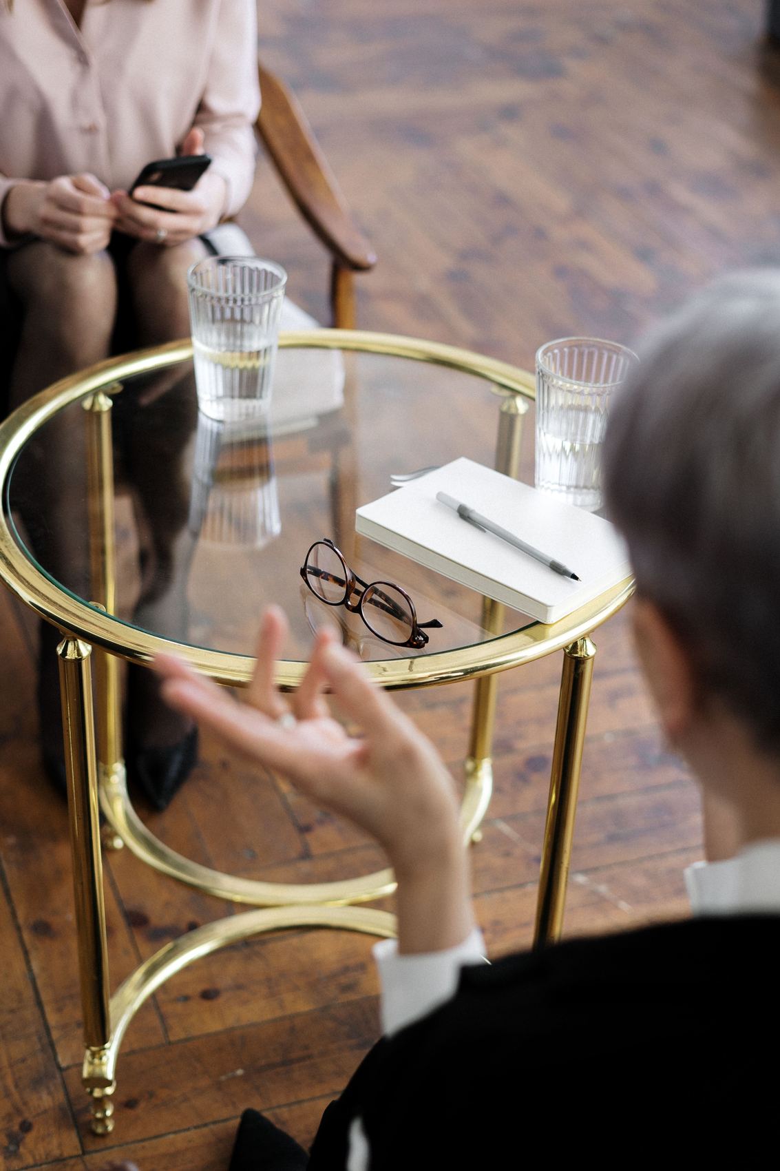 Two people talking across a glass table set with water, a notebook and a pair of glasses